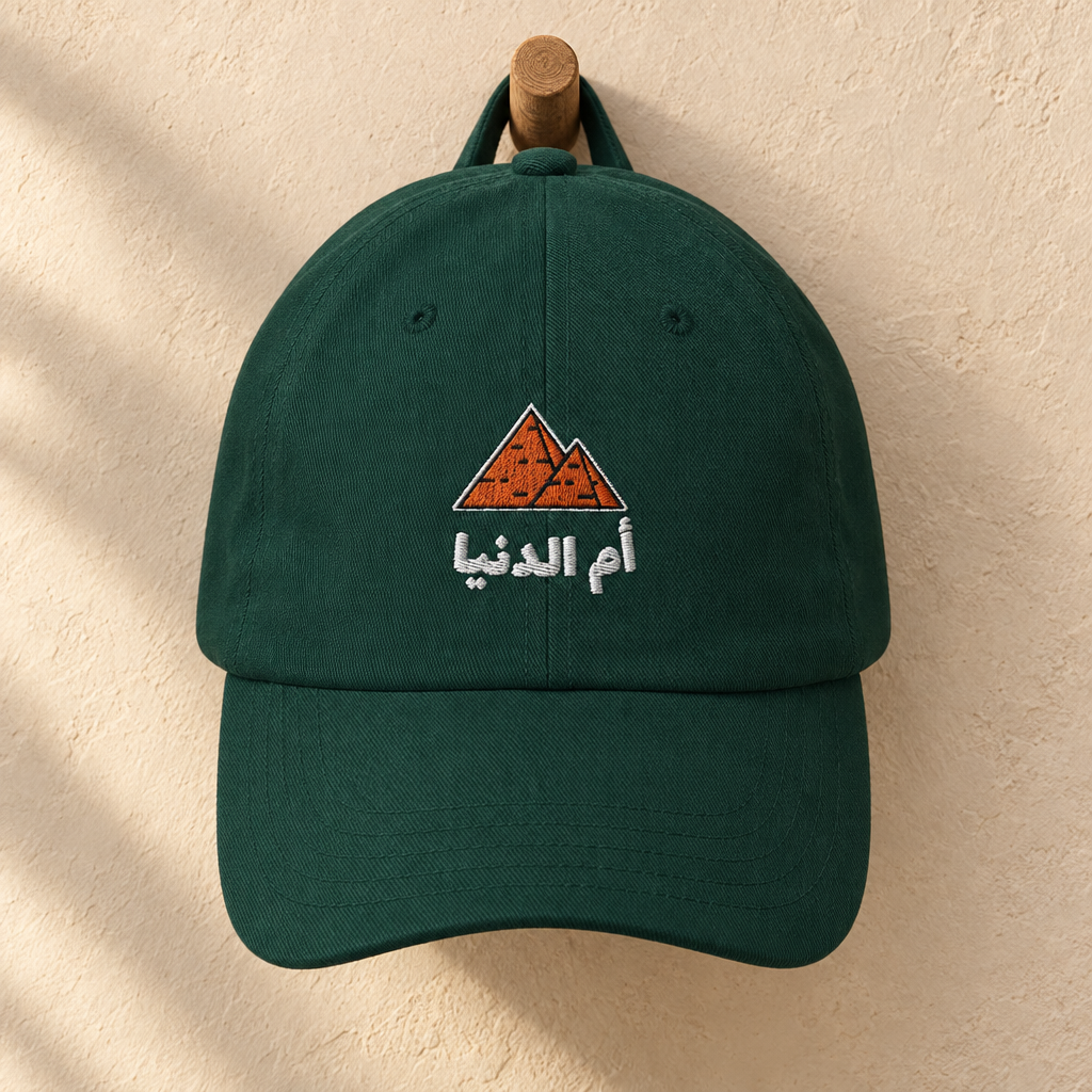 Spruce green Egypt Umm El Dunya cap with embroidered pyramids and Arabic lettering, hanging on a wooden peg.