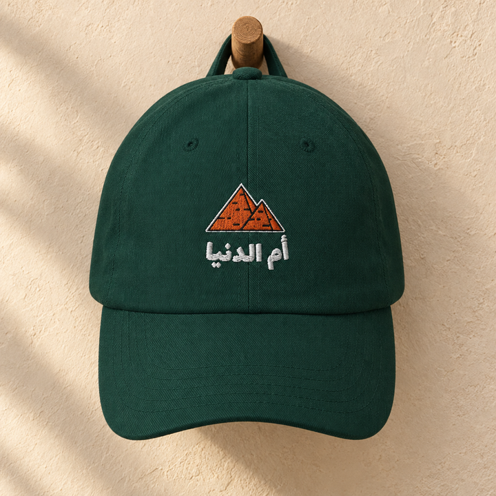 Spruce green Egypt Umm El Dunya cap with embroidered pyramids and Arabic lettering, hanging on a wooden peg.