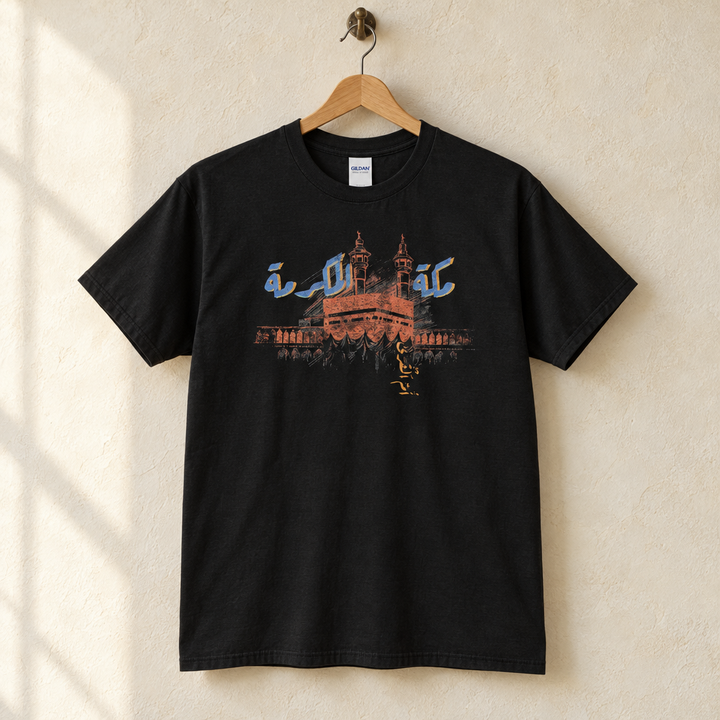 Makkah Al Mukarramah T-shirt, hanging on a wooden hanger against a pale wall.